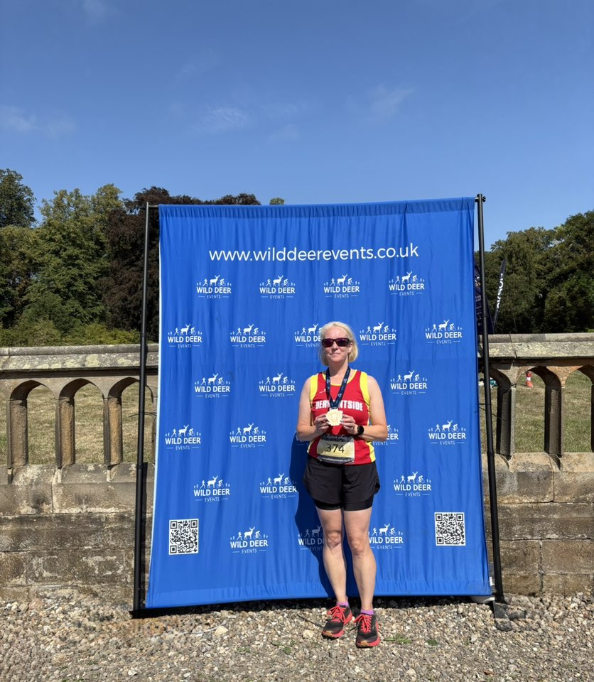 A picture of Emma proudly holding her medal after a race. She is standing in front of a Wild Deer Events banner.