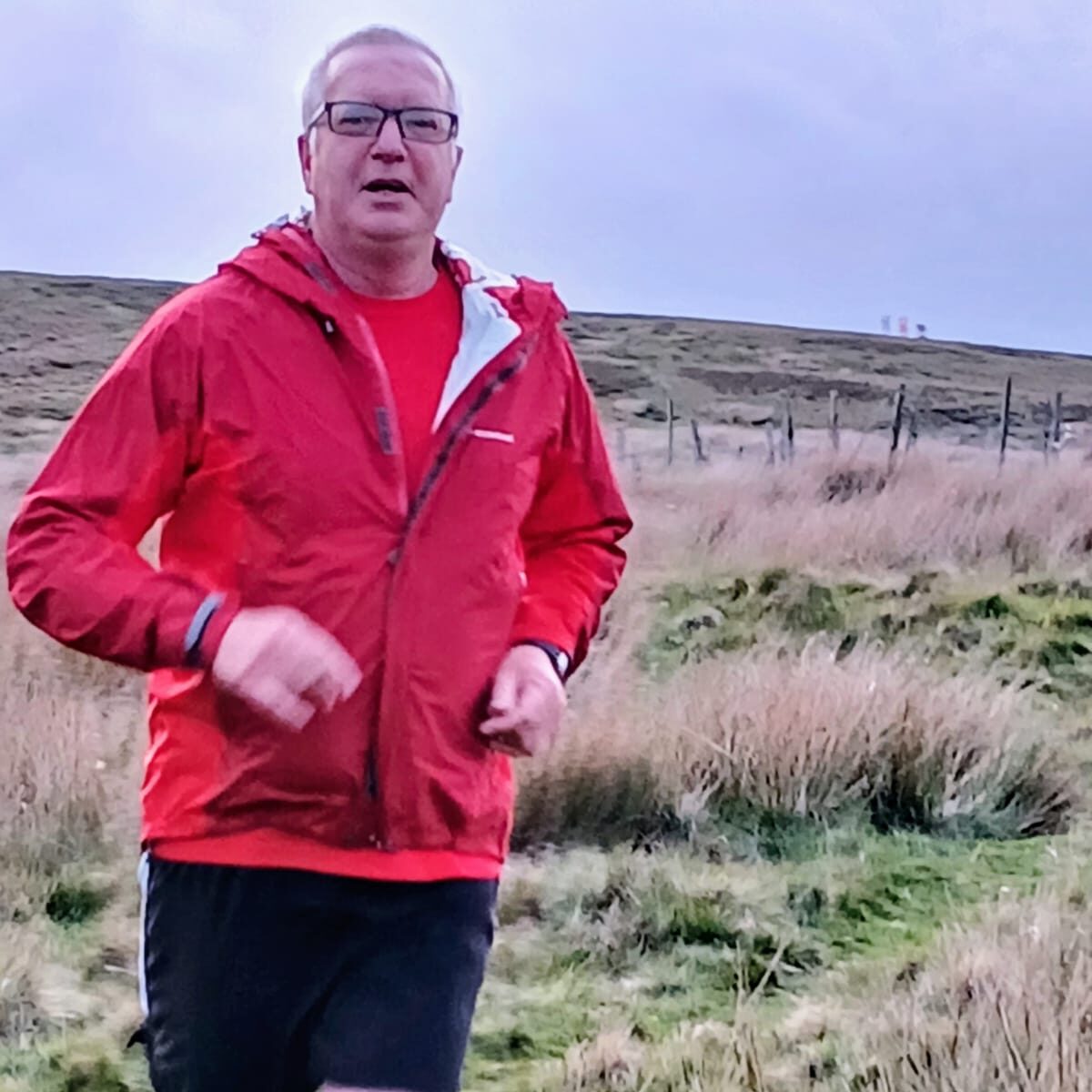 A picture of David Howe on a run in the countryside. He is wearing a red running jacket, black shorts and glasses.